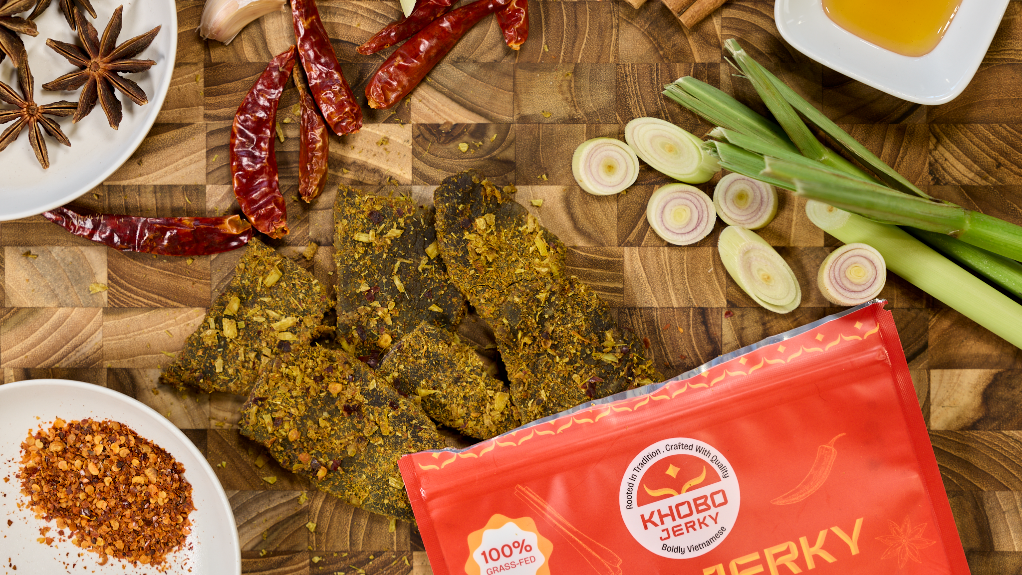 handcrafted grass fed vietnamese jerky displayed with fresh ingredients, lemongrass, dried chili flakes, whole spices like star anise, cinnamon and honey on a wooden board